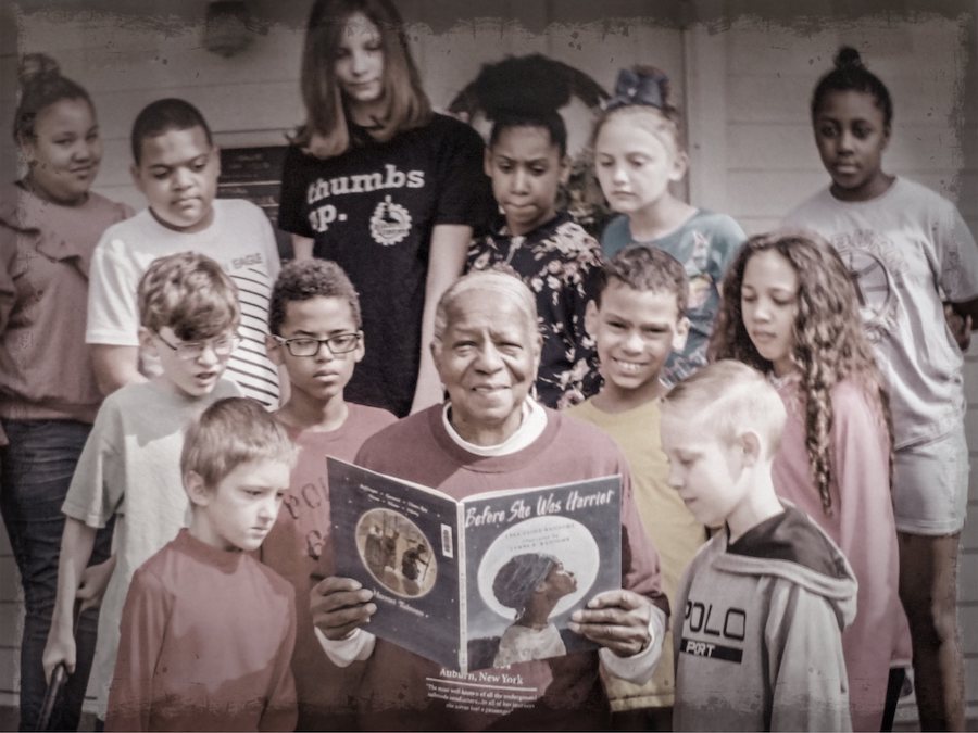 photo of Ms. Copes Johnson holding a book open, surrounded by 10 young students