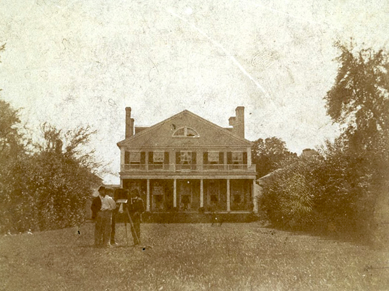 grainy, sepia-toned photograph of the regal front facade of a plantation house, surrounded by low trees